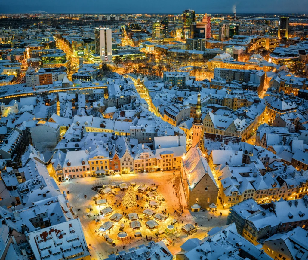 An aerial view of a snow-covered European city at night, with a brightly lit central square featuring a large Christmas tree and market stalls.