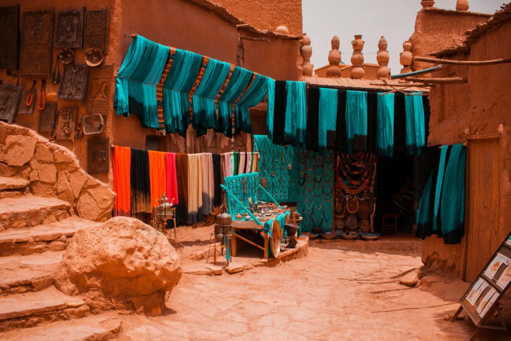A vibrant street scene in a traditional Moroccan town, with a stall selling colorful textiles and crafts. Blue and multi-colored fabrics hang from awnings and lines, and an old stone staircase is visible on the left.