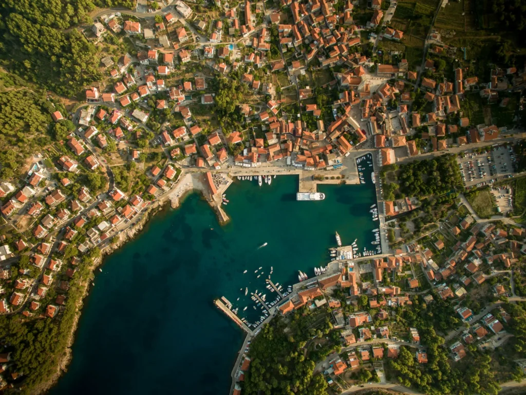 An aerial perspective of a densely built coastal town with red roofs, nestled into a hillside overlooking a calm, blue harbor with boats.