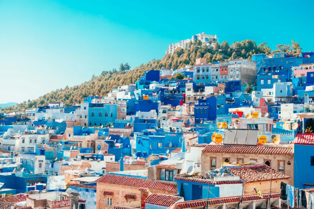 A panoramic view of the blue-painted buildings of Chefchaouen, Morocco, clinging to a hillside. A fortress or kasbah is visible at the top of the hill, surrounded by trees, under a bright blue sky.