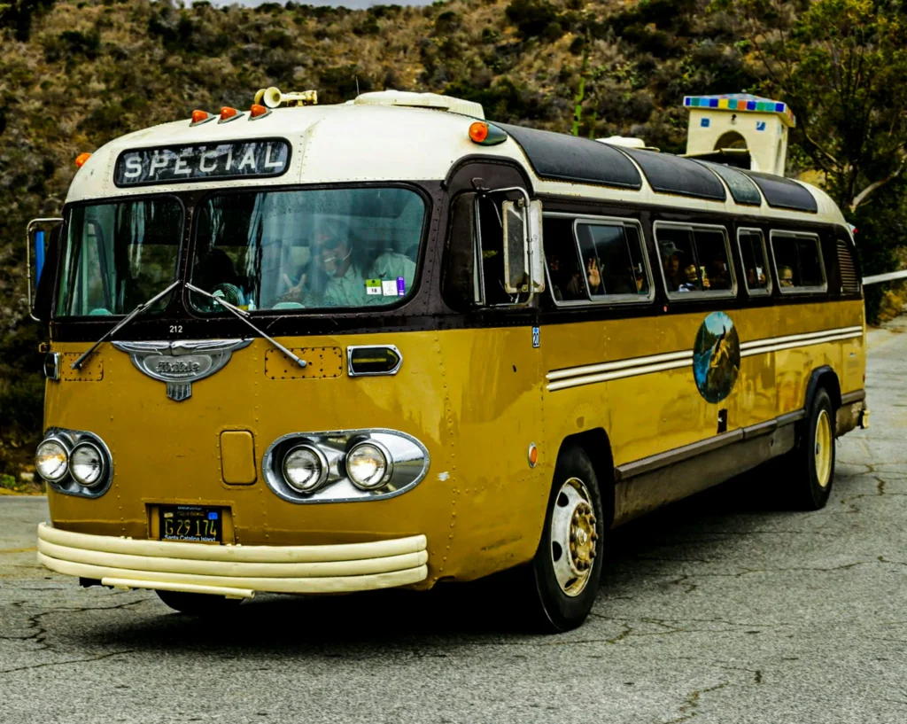 A vintage brown and cream-colored bus with "SPECIAL" on its destination sign and a decorative horn on the roof is parked on an asphalt surface, with a rocky hillside in the background.