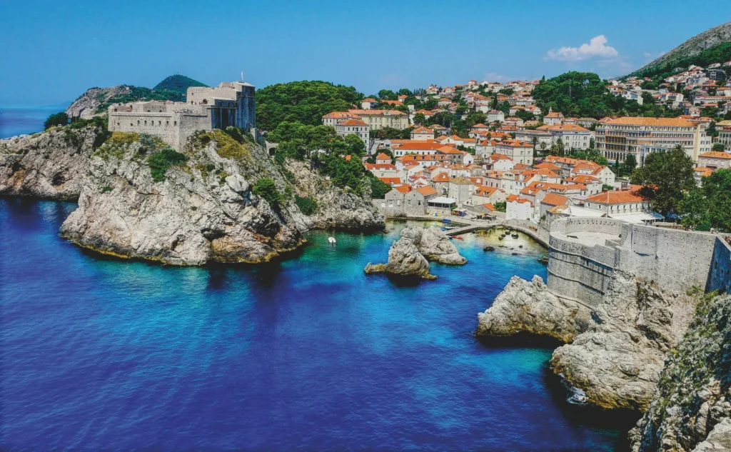 A picturesque coastal town featuring red-tiled roofs and ancient stone fortifications, set against a rugged blue coastline and deep blue sea.
