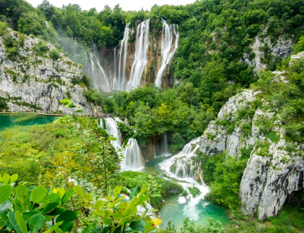 Multiple levels of vibrant green waterfalls cascading into turquoise lakes within a lush, forested national park (likely Plitvice Lakes, Croatia).