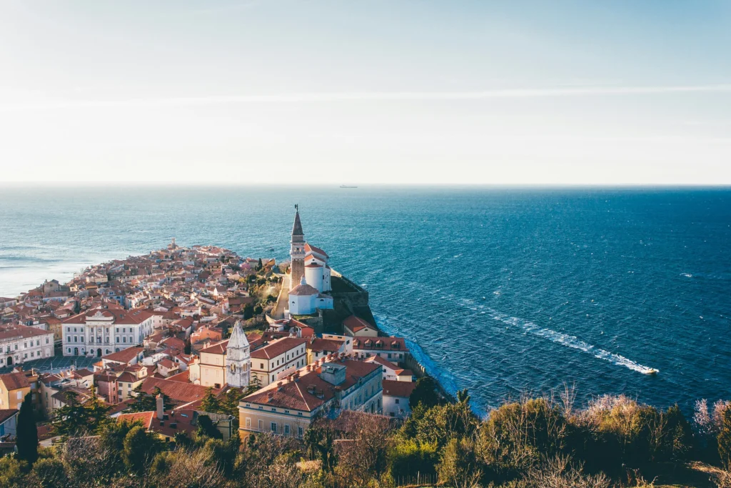 An elevated view captures the historic coastal town of Piran, Slovenia, jutting into the clear blue Adriatic Sea. The town is densely packed with terracotta-roofed buildings and features a prominent church with a tall bell tower at its highest point. The sea stretches to the horizon, with a boat's wake visible in the water.