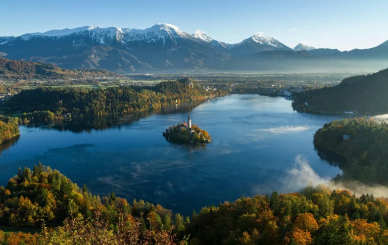 An aerial view of Lake Bled, Slovenia, featuring a small island with a church, surrounded by colorful autumn foliage and distant mountains under a clear sky.
