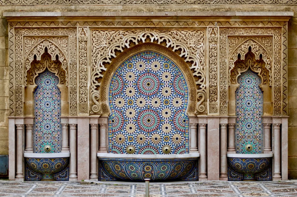 An intricately decorated Moroccan mosaic fountain with an arched central panel and two smaller arched side panels, all adorned with colorful geometric patterns and surrounded by carved stone.