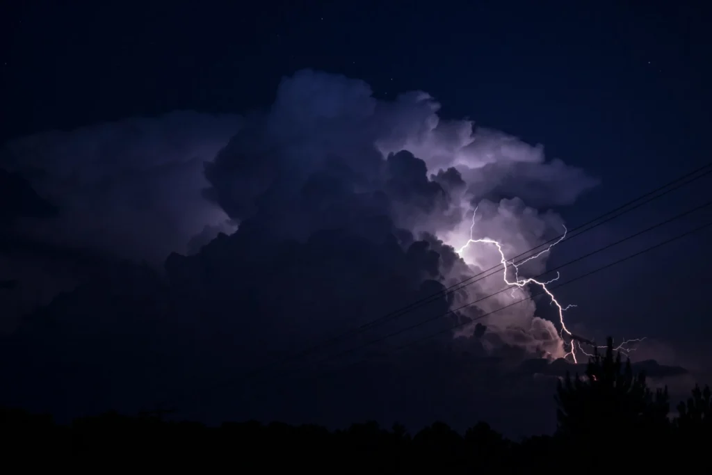A powerful lightning bolt illuminates dark, turbulent storm clouds over a silhouetted tree line at night.