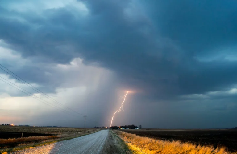 A long, wet road stretches into the distance under a dramatic stormy sky, with a bright lightning strike hitting the ground in the mid-ground.