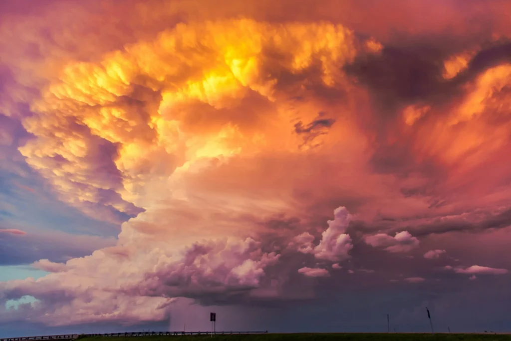 A vast, fiery sky filled with enormous, swirling clouds in vibrant shades of orange, red, and purple, suggesting a dramatic sunset or sunrise over a dark landscape.