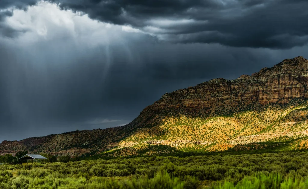 Streaks of rain fall from dark storm clouds over a partially sunlit hillside of green and yellow vegetation, creating a moody landscape.