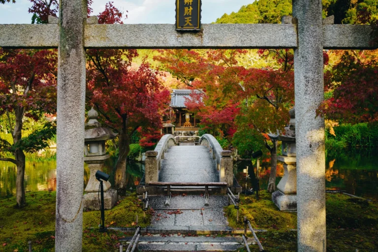 A stone torii gate in a Japanese garden setting, with red and orange autumn foliage surrounding a stone bridge leading to a traditional building in the background.