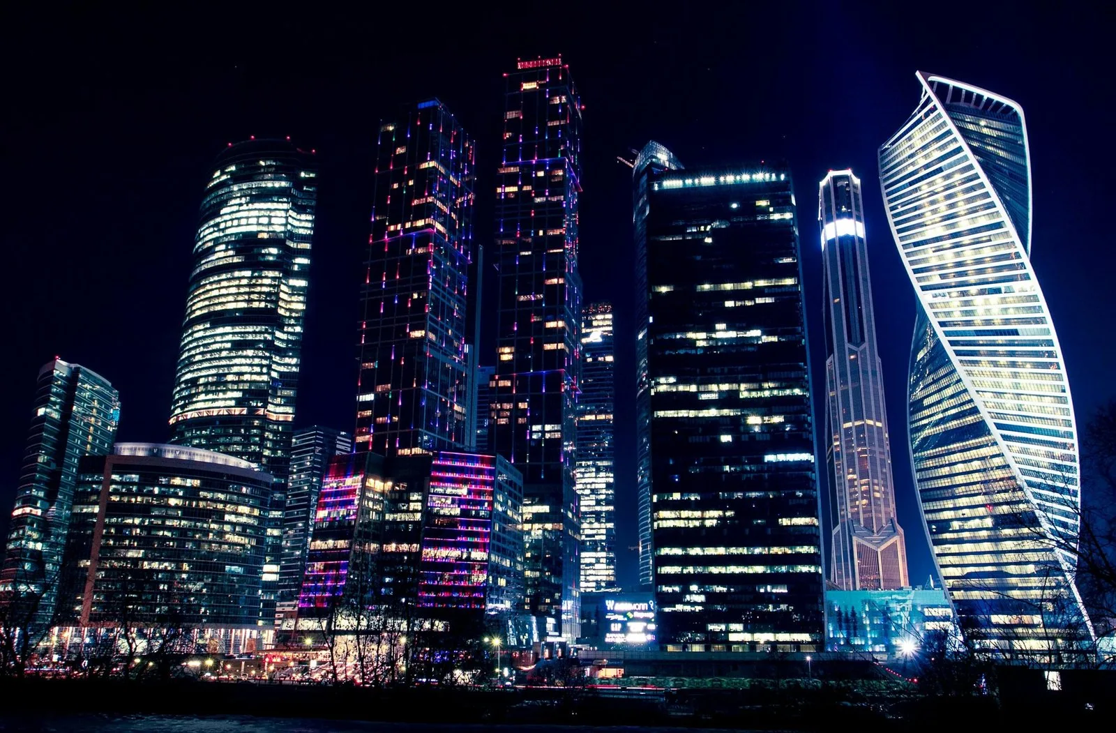 A nighttime view of a cluster of modern, illuminated skyscrapers reflecting lights.