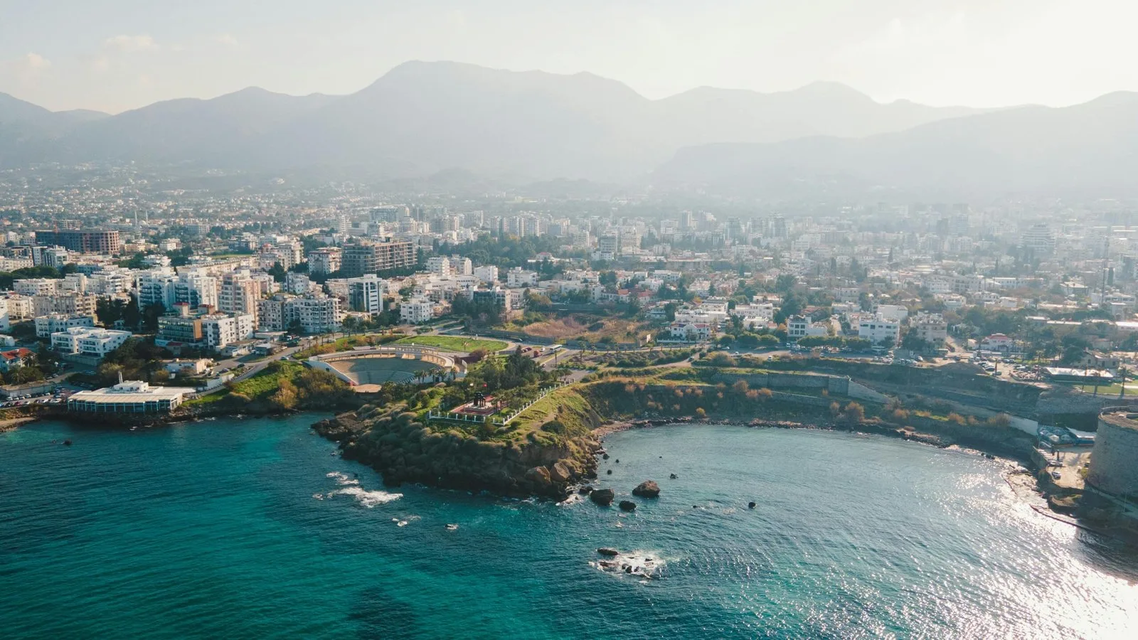 This image captures a scenic cove with a city skyline in the background. The location is Ayia Napa, specifically showcasing one of its beautiful coastal areas and pristine waters. Ayia Napa is a well-known resort town on the southeastern coast of Cyprus, famous for its beaches and nightlife.