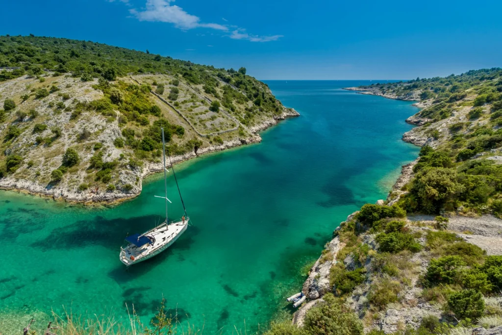 A serene, secluded turquoise bay with a small white boat anchored in crystal-clear water, surrounded by green hills and a rocky coastline under a clear blue sky.