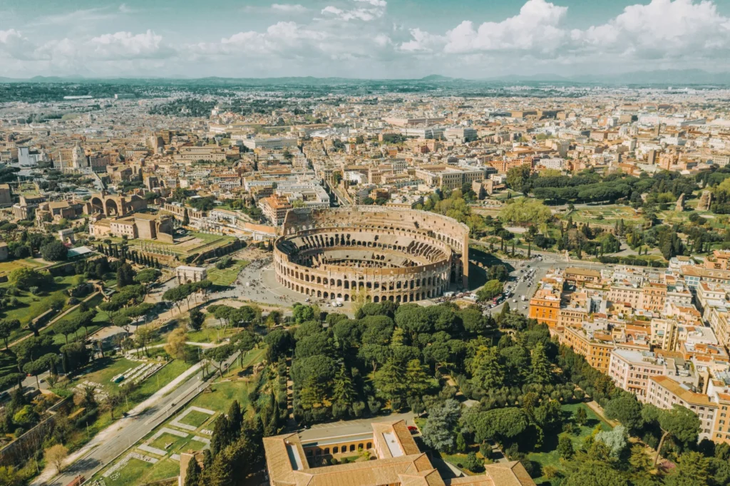 Aerial or high-angle view of Rome, featuring the Colosseum in the mid-ground surrounded by green space, and the expansive urban landscape beyond under a cloudy sky.
