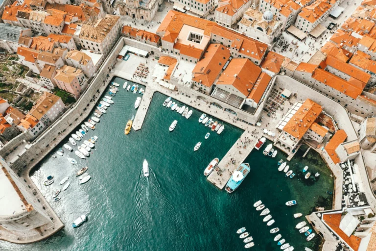 An aerial view of a vibrant European harbor filled with white boats, surrounded by a historic town with numerous red-tiled rooftops.