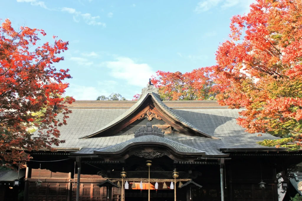 A traditional Japanese temple or shrine building with an ornate roof is framed by vivid red autumn maple trees against a blue sky.
