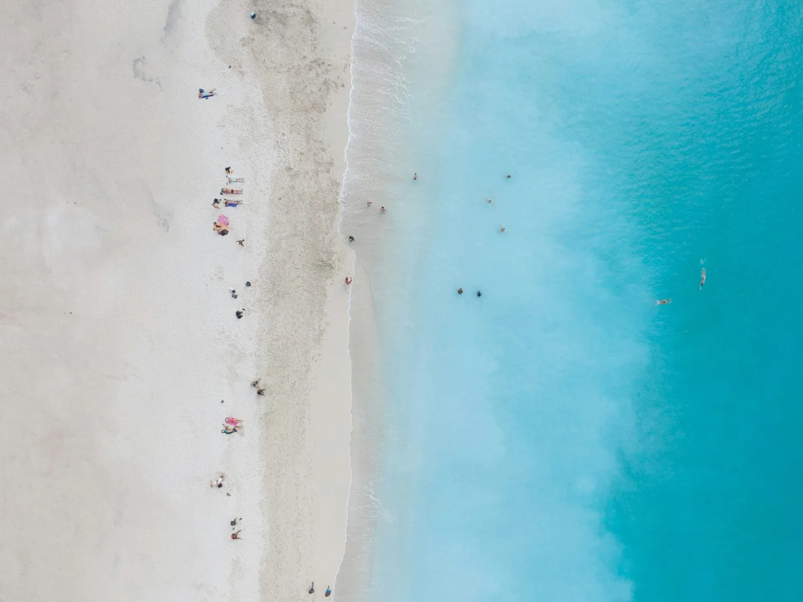 An aerial view of a white-sand beach and turquoise water, with people and umbrellas on the shore.