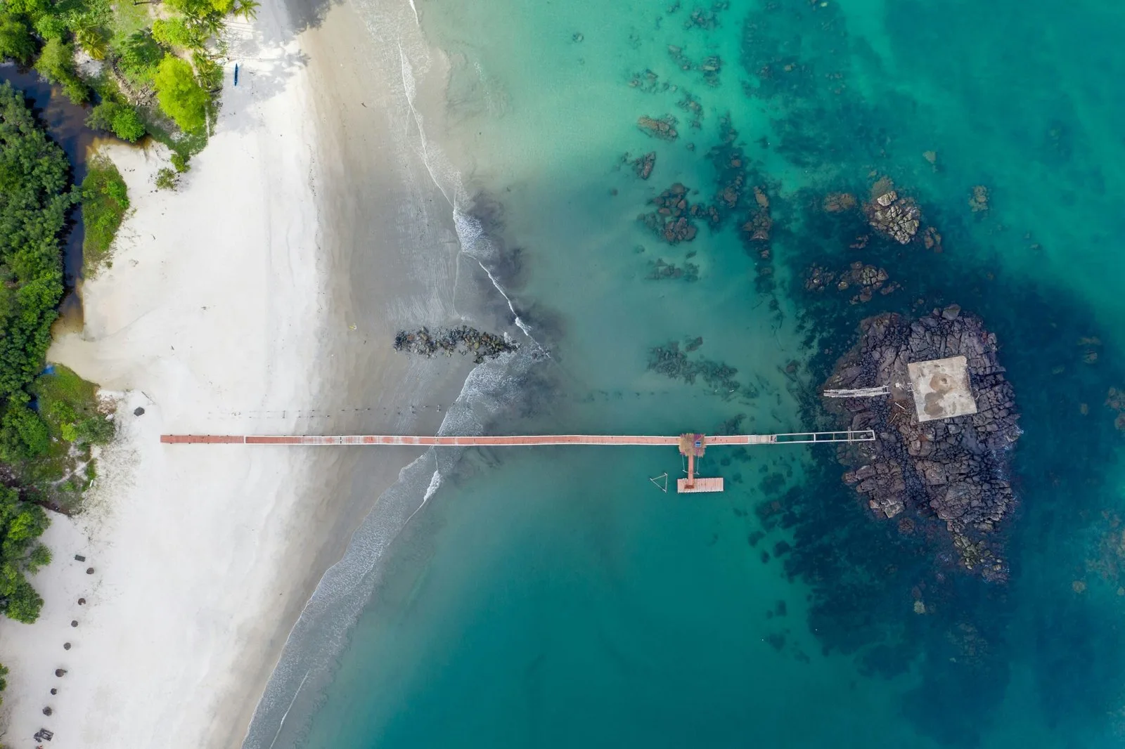 An aerial shot of a small beach with a wooden pier extending into the clear, blue water.