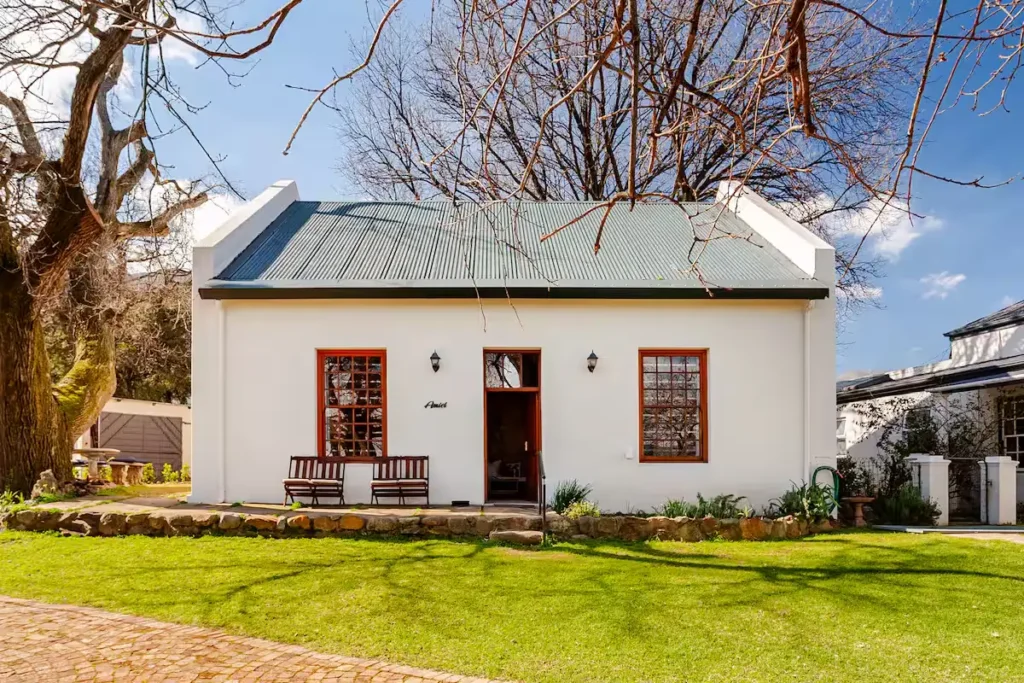 Off-season white country cottage with empty benches under leafless trees and vibrant green lawn, a quiet travel retreat.