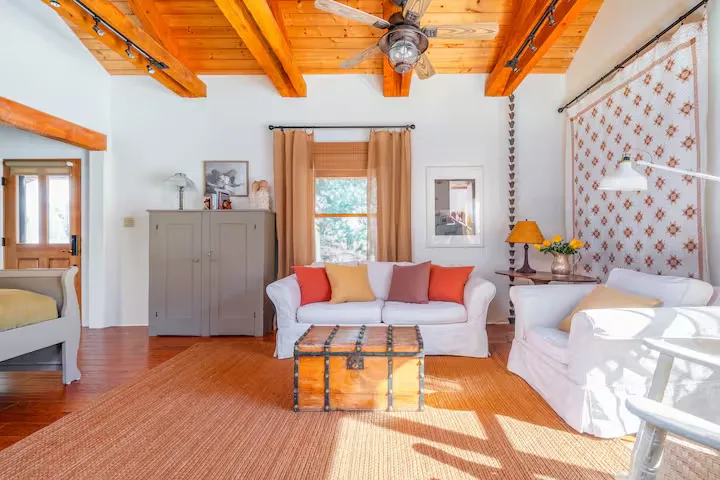 Cozy sunlit vacation rental living room with white sofas, colorful pillows, exposed beams and trunk table, off-season calm.