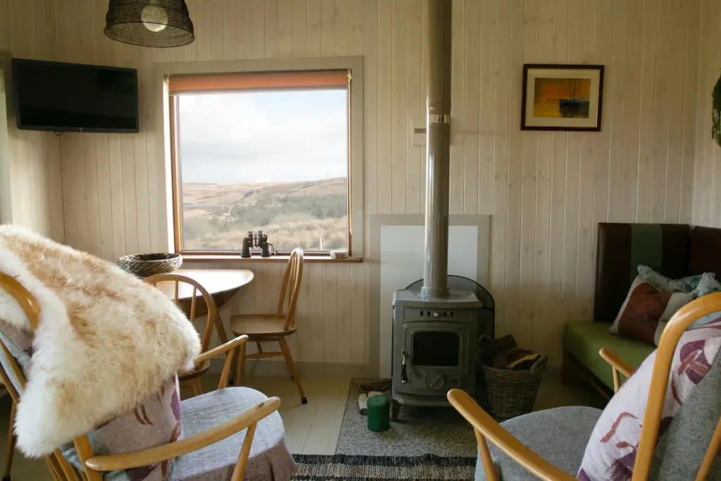 Cozy off-season travel cabin retreat interior with wood-burning stove, fur throws and empty chairs by misty hillside window