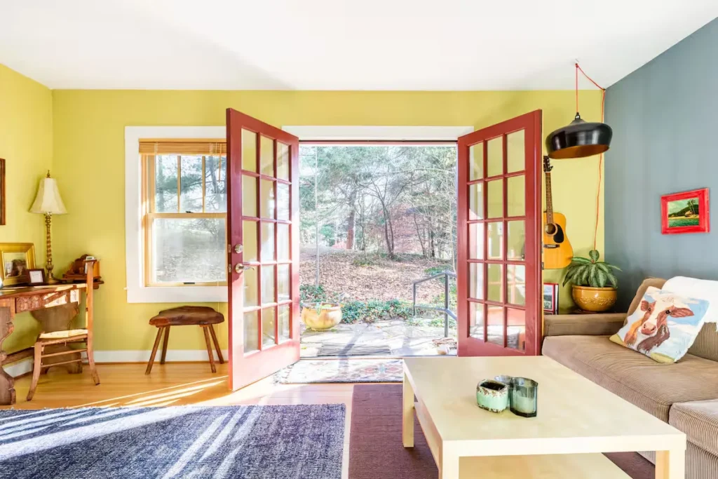 Bright cozy living room with open red French doors revealing a leaf-strewn patio and tranquil off-season woods beyond.