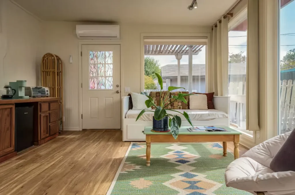 Bright off-season vacation rental studio interior with cozy daybed, coffee table plant, wood floors and sunlit windows.