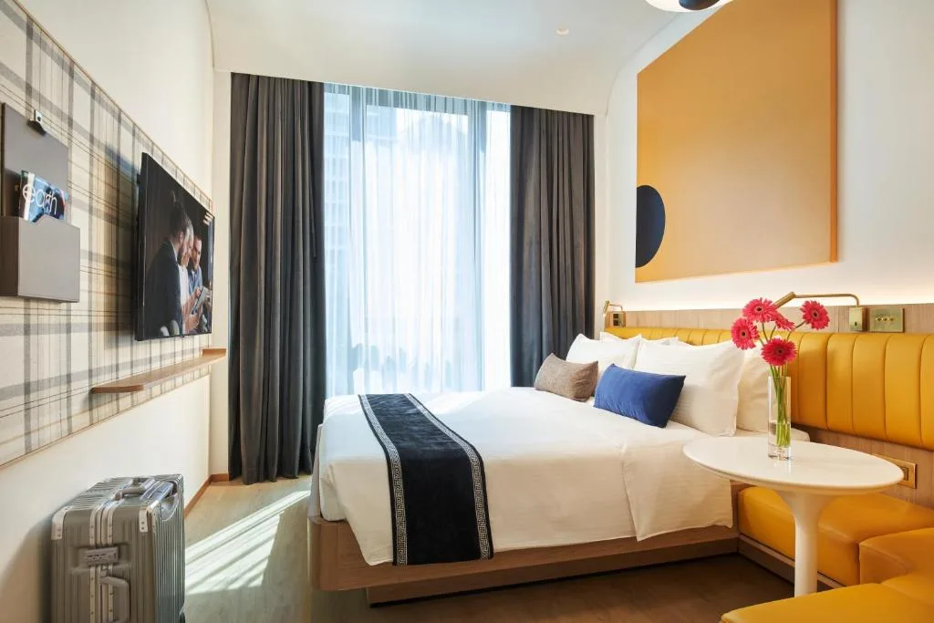 Bright modern hotel room with queen bed, yellow banquette seating, fresh flowers and lone suitcase, off-season quiet retreat