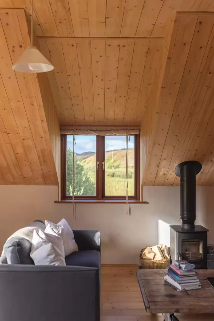 Cozy cabin loft living room with wood stove, soft sofa and books, sunlit window framing a quiet off-season hillside view