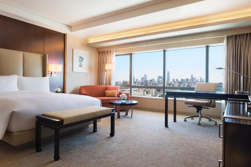 Modern hotel room with king bed, sofa and desk, wide city skyline view through large windows, evoking tranquil off-season travel vibe.