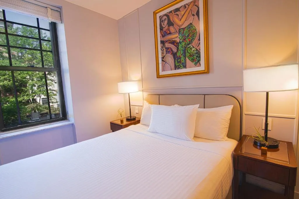 Cozy hotel room with queen bed, bedside lamps and leafy window view, a serene off-season travel retreat.