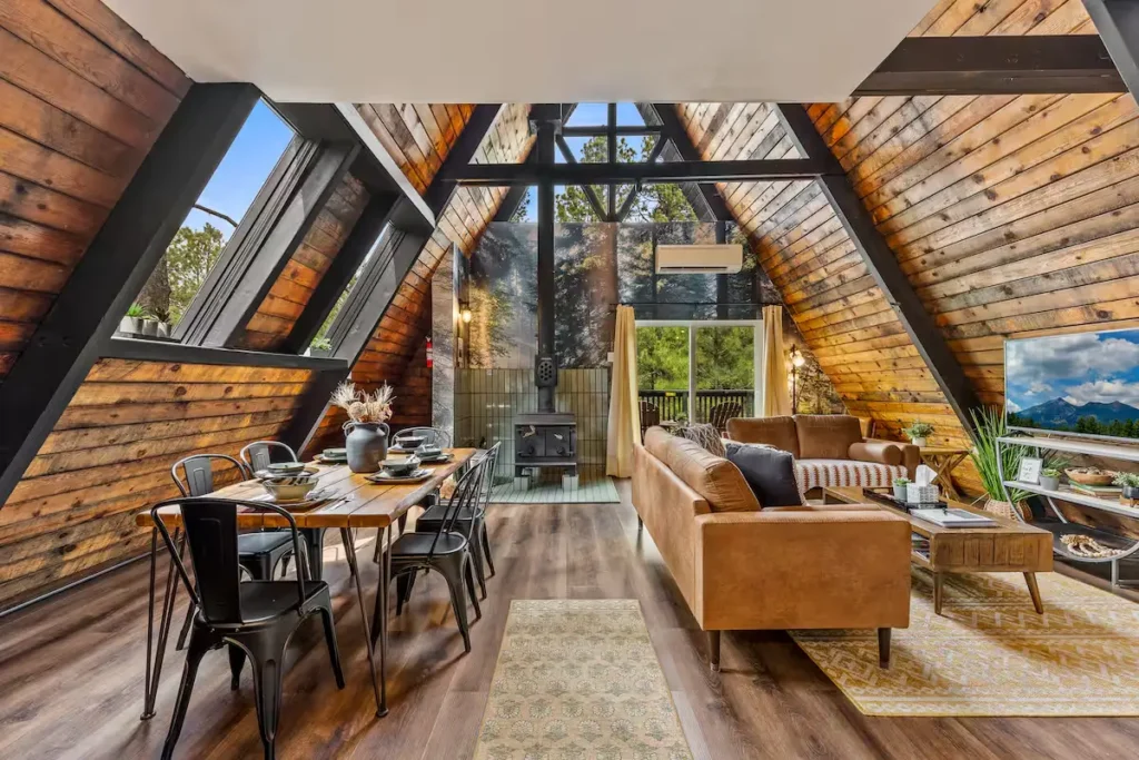 Off-season A-frame cabin interior with wood stove, cozy leather couch, dining table and windows overlooking forest trees