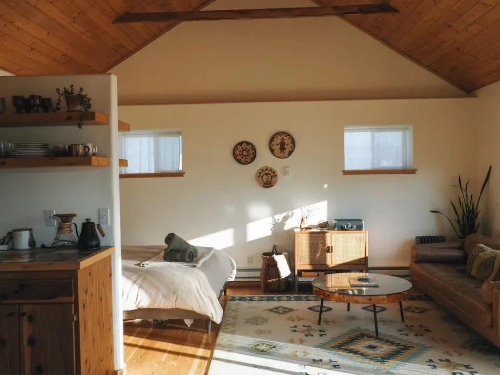 Sunlit boho loft studio cabin interior with cozy bed and minimalist kitchen, an off-season remote retreat.