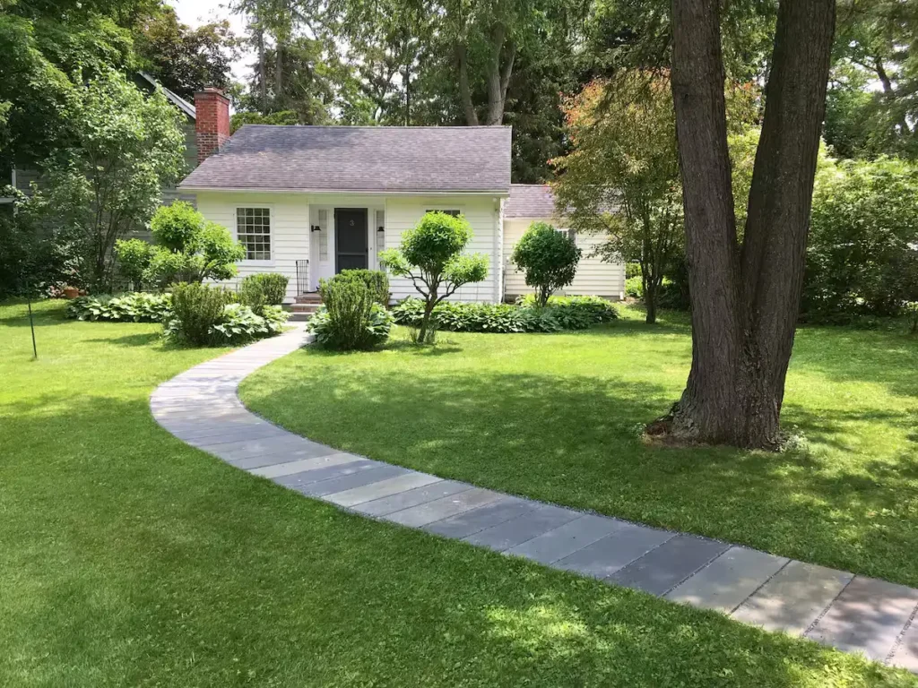 Serene off-season travel cottage with curved stone path, lush green lawn and surrounding trees evoking quiet retreat charm.