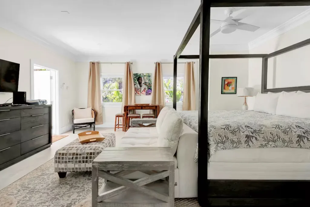 Bright airy beachside suite with four-poster bed, sofa and table overlooking lush palms, serene off-season ambience.