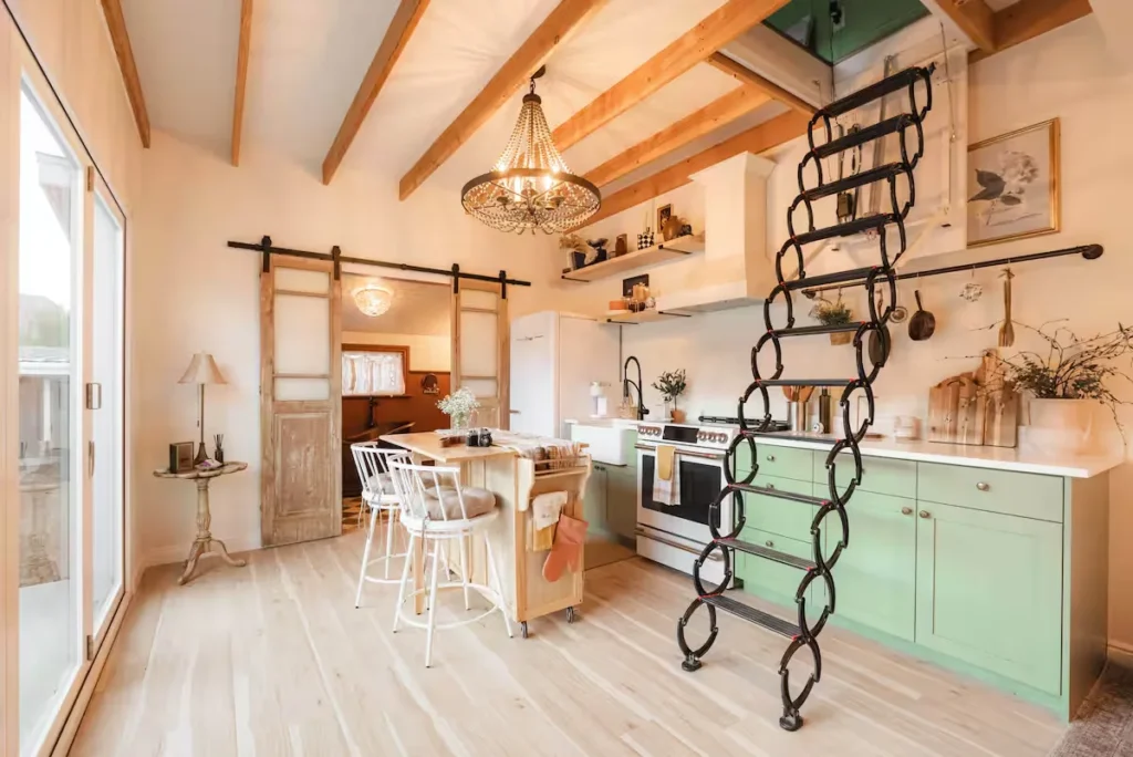 Sunlit off-season cabin kitchen with exposed beams, green cabinetry, retractable loft ladder and sliding barn doors.