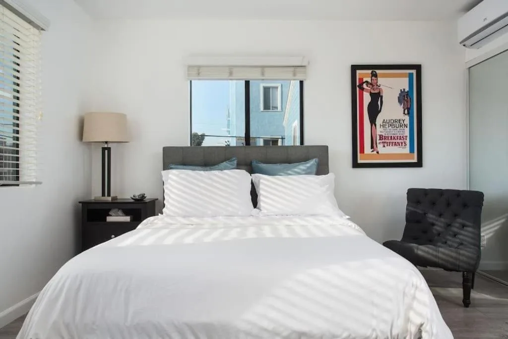 Bright minimalist vacation bedroom with white linens, natural light streaming through blinds, a calm off-season retreat.