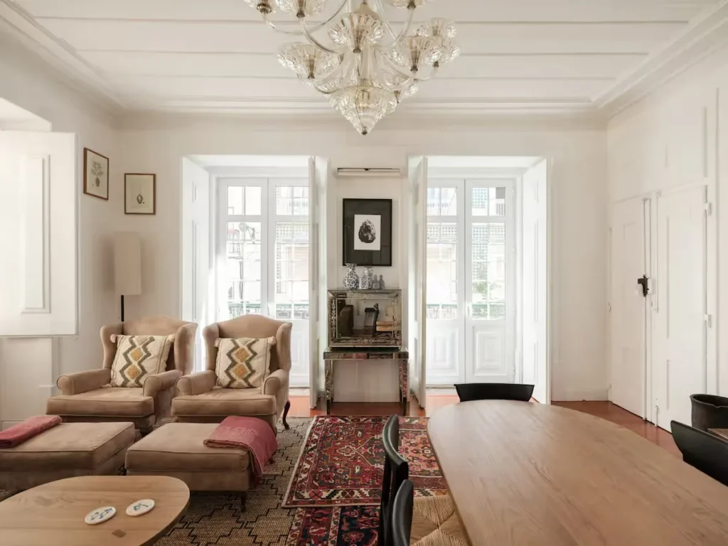 Sunlit off-season vacation rental living room with plush armchairs, chandelier, hardwood floors and open white shutters.