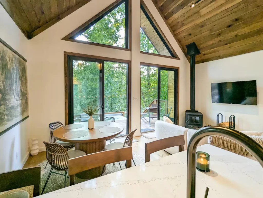 Cozy off-season forest cabin interior with vaulted wood ceiling, dining table, living area and deck overlooking empty woods.