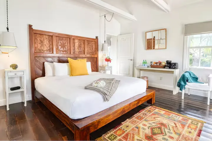 Bright boutique hotel room with carved wooden headboard, colorful rug and tropical glow, quiet off-season beach retreat vibe
