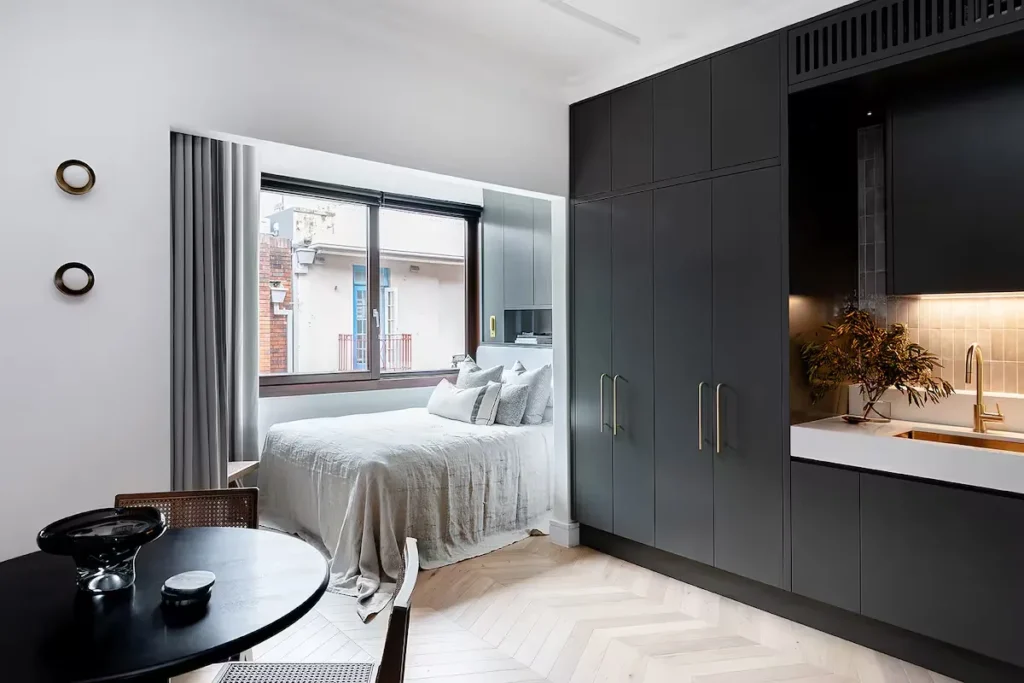 Minimalist studio apartment with bed by window, dark kitchenette and small table, creating a quiet off-season travel retreat.