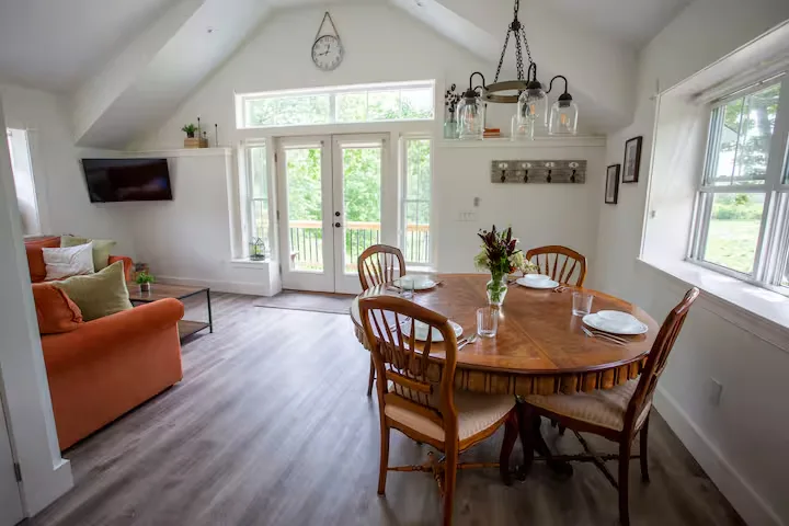 Airy off-season cabin vacation rental dining area with round wooden table, cozy living space and leafy forest-view deck.