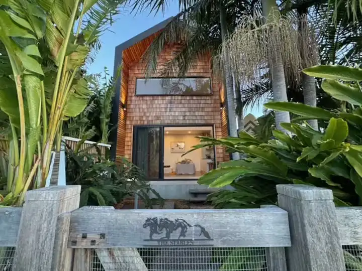 Cozy wood-shingle tropical cabin nestled among palm trees behind a weathered fence, off-season quiet vacation rental vibe