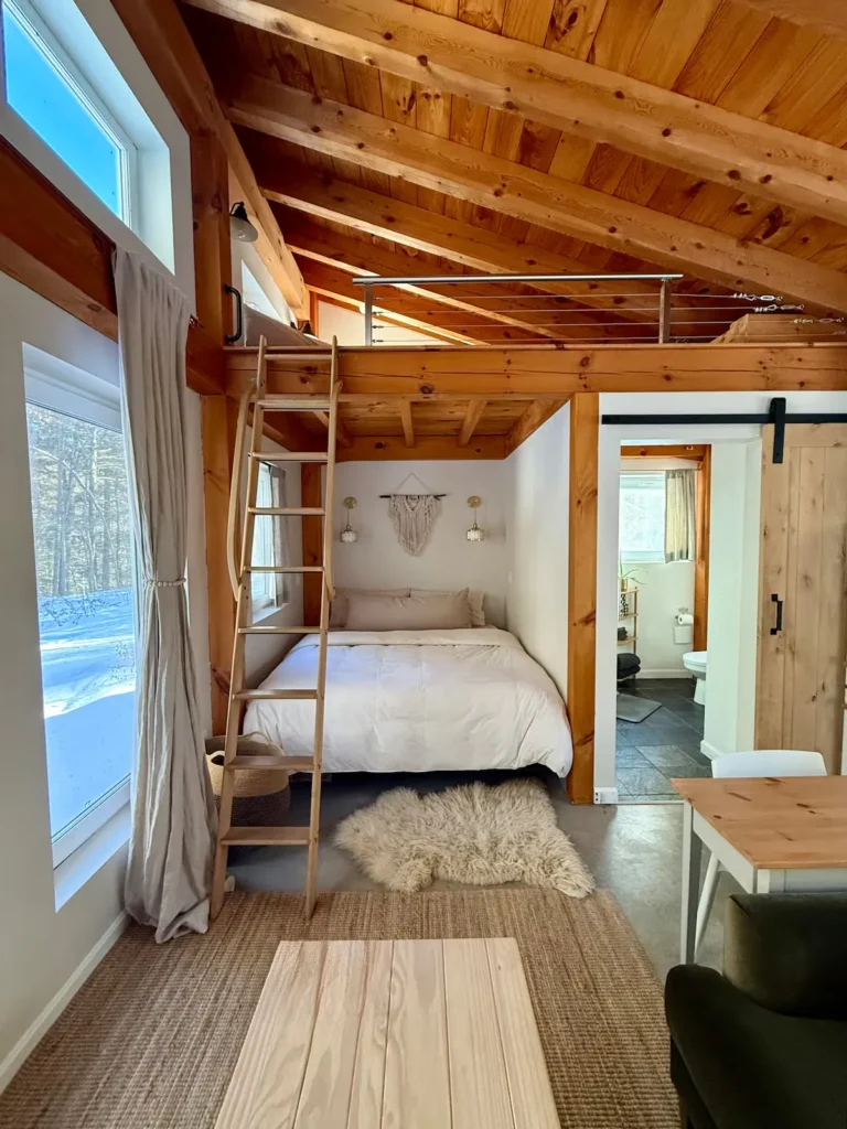 Cozy winter cabin interior with loft bed, rustic wood beams and large windows overlooking a snowy forest off-season retreat.