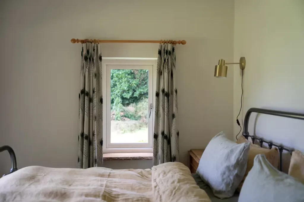 Cozy off-season cottage bedroom with iron bed, neutral linens and a leafy garden view through the window.