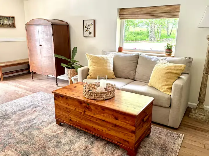 Cozy off-season cottage living room with beige sofa, yellow pillows, rustic wood table, plush rug and quiet green window view