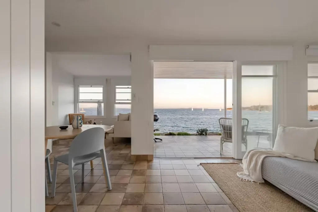 Sunlit, minimalist beach house interior opening onto a quiet off-season seaside terrace with sailboats.