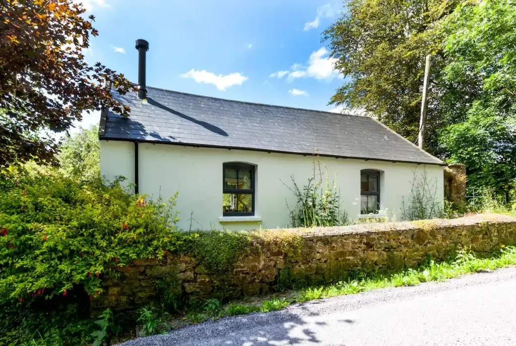Secluded white countryside cottage beside a mossy stone wall on a quiet off-season roadside, lush greenery.