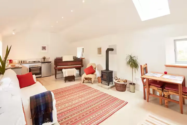 Cozy off-season cottage interior with wood-burning stove, piano, dining nook and patterned rug brightly lit by skylight.